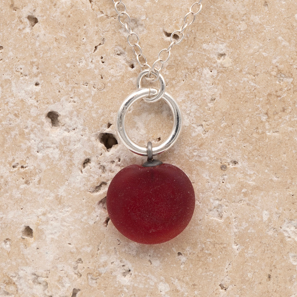 Close up of the back of a red frosted glass charm with swirl imprint on a sandstone background. The whole charm is frosted, and spiral is plain frosted glass. The charm hangs on a large silver jump ring. The necklace is completed with a small silver jump ring and silver chain.