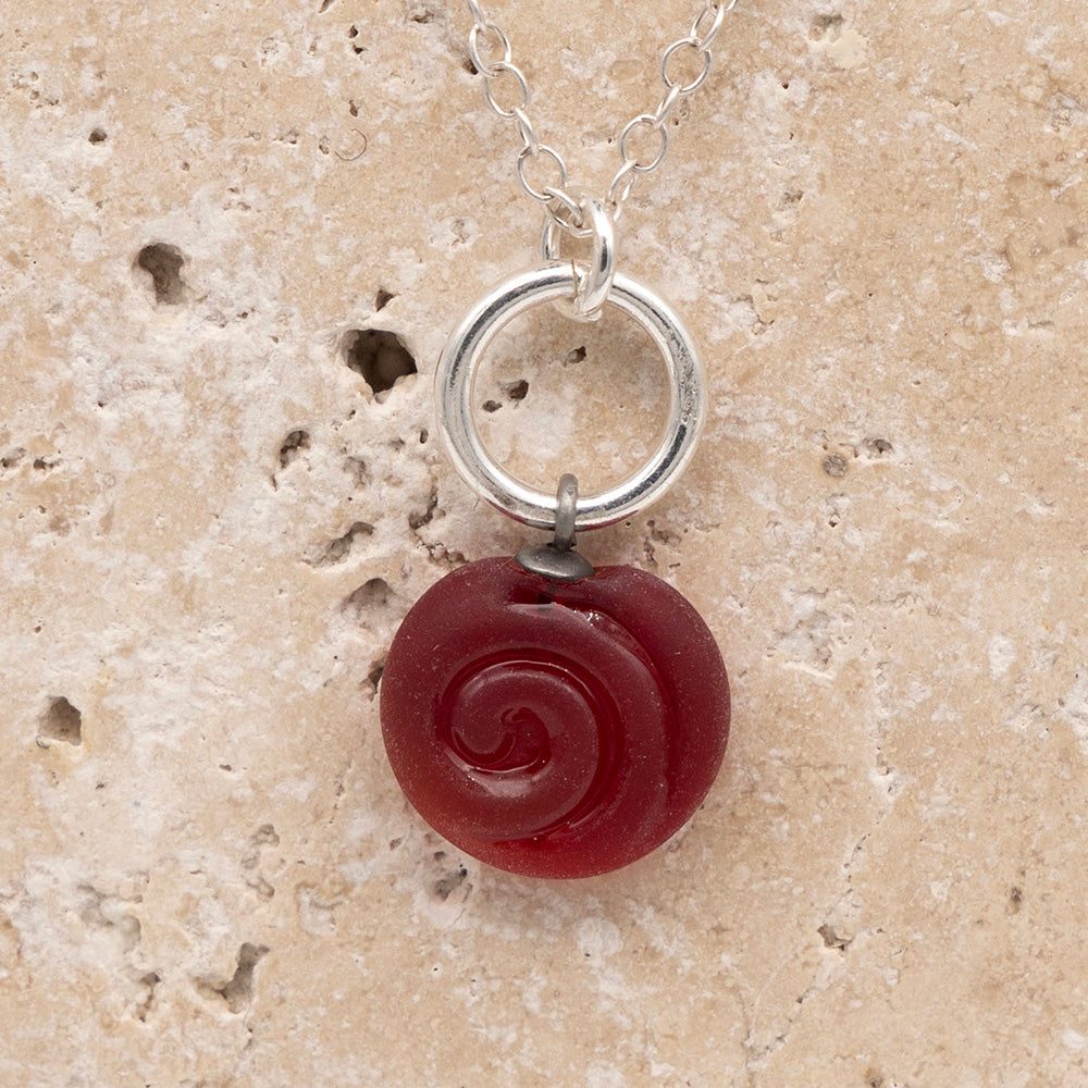 Close up of the front  of a red frosted glass charm with swirl imprint on a sandstone background. The charm is frosted and spiral is shiny. The charm hangs on a large silver jump ring. The necklace is completed with a small silver jump ring and silver chain.