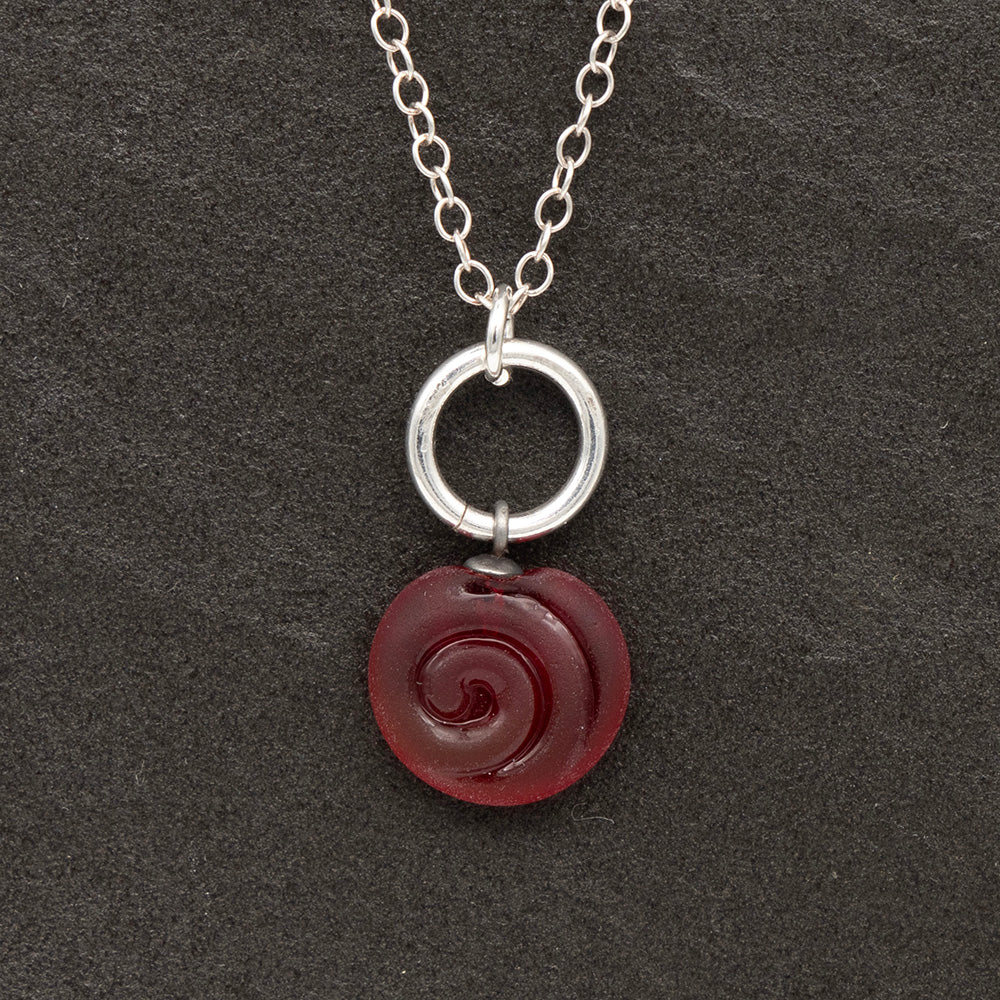 Close up of red frosted glass charm with swirl imprint on a dark slate background. The charm is frosted and spiral is shiny. The charm hangs on a large silver jump ring. The necklace is completed with a small silver jump ring and silver chain.