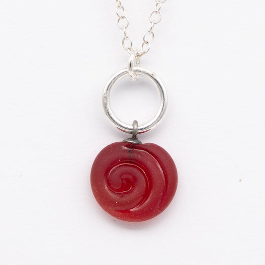 Close up of red frosted glass charm with swirl imprint. The charm is frosted and spiral is shiny. The charm hangs on a large silver jump ring. The necklace is completed with a small silver jump ring and silver chain.