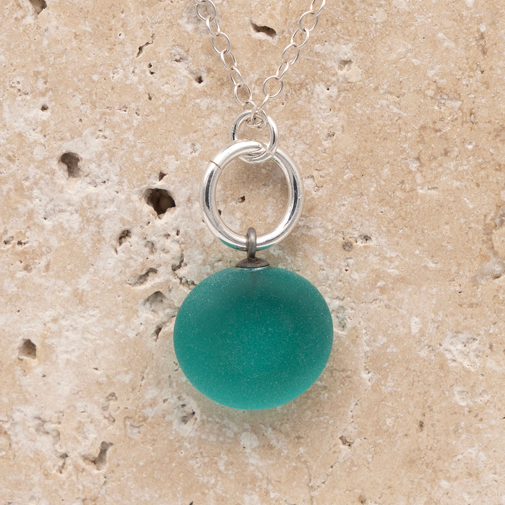 Close up of the back of a teal frosted glass charm with swirl imprint on a sandstone background. The whole charm is frosted, and spiral is plain frosted glass. The charm hangs on a large silver jump ring. The necklace is completed with a small silver jump ring and silver chain.