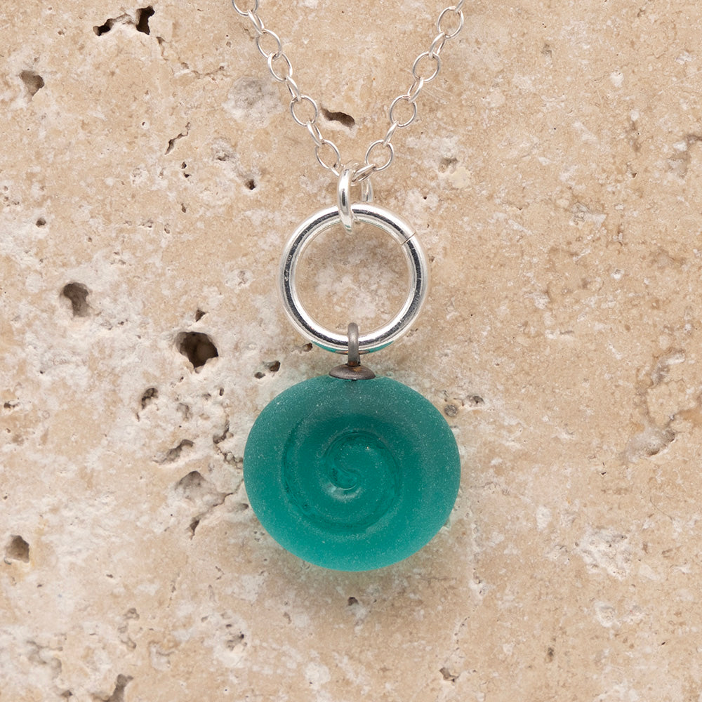 Close up of the front  of a teal frosted glass charm with swirl imprint on a sandstone background. The charm is frosted and spiral is shiny. The charm hangs on a large silver jump ring. The necklace is completed with a small silver jump ring and silver chain.