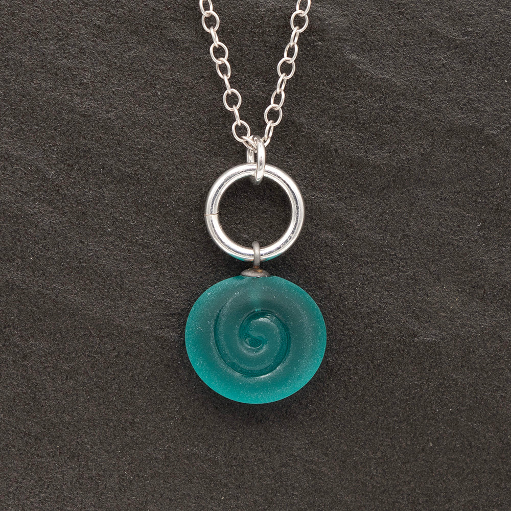 Close up of a teal frosted glass charm with swirl imprint on a dark slate background. The charm is frosted and spiral is shiny. The charm hangs on a large silver jump ring. The necklace is completed with a small silver jump ring and silver chain.