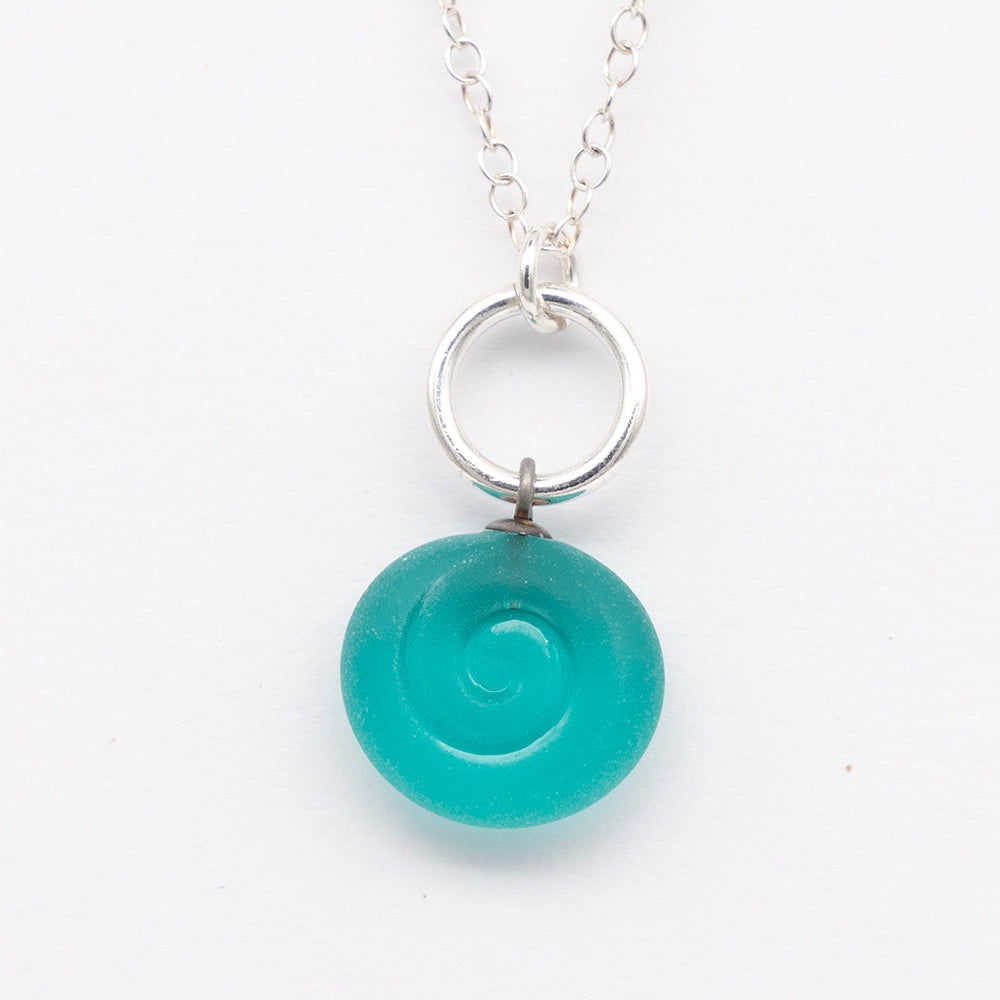 Close up of teal frosted glass charm with swirl imprint. The charm is frosted and spiral is shiny. The charm hangs on a large silver jump ring. The necklace is completed with a small silver jump ring and silver chain.