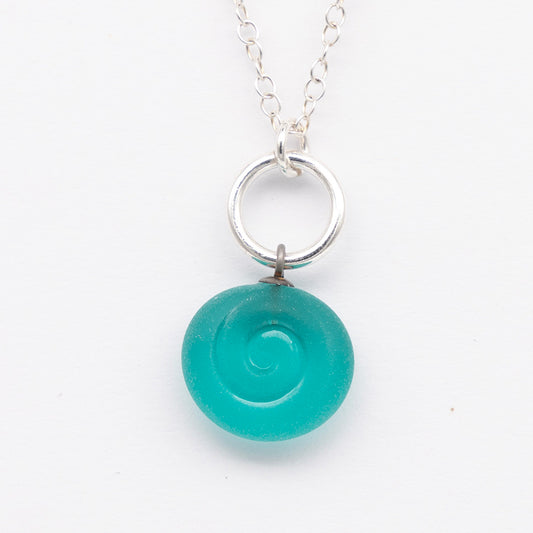 Close up of teal frosted glass charm with swirl imprint. The charm is frosted and spiral is shiny. The charm hangs on a large silver jump ring. The necklace is completed with a small silver jump ring and silver chain.