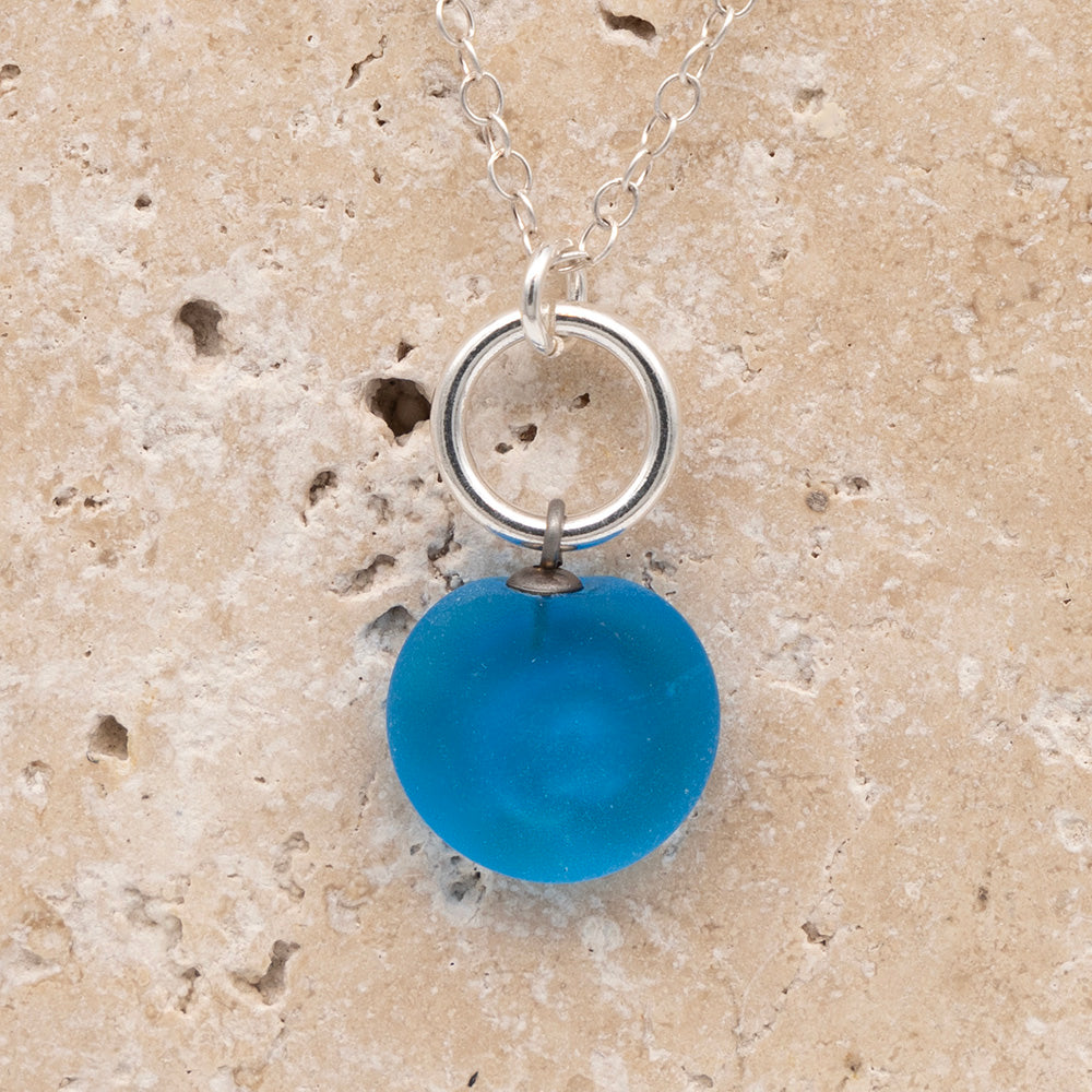 Close up of the back of a turquoise frosted glass charm with swirl imprint on a sandstone background. The whole charm is frosted, and spiral is plain frosted glass. The charm hangs on a large silver jump ring. The necklace is completed with a small silver jump ring and silver chain.