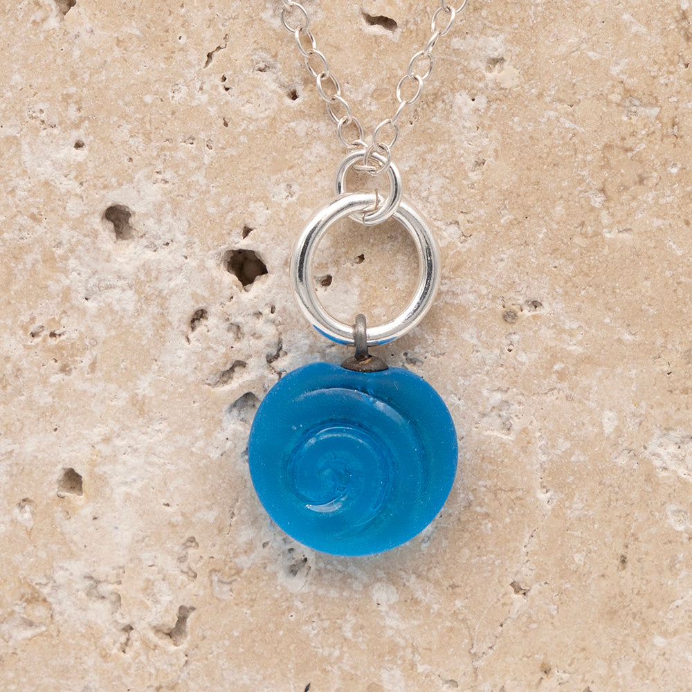 Close up of the front  of a turquoise frosted glass charm with swirl imprint on a sandstone background. The charm is frosted and spiral is shiny. The charm hangs on a large silver jump ring. The necklace is completed with a small silver jump ring and silver chain.