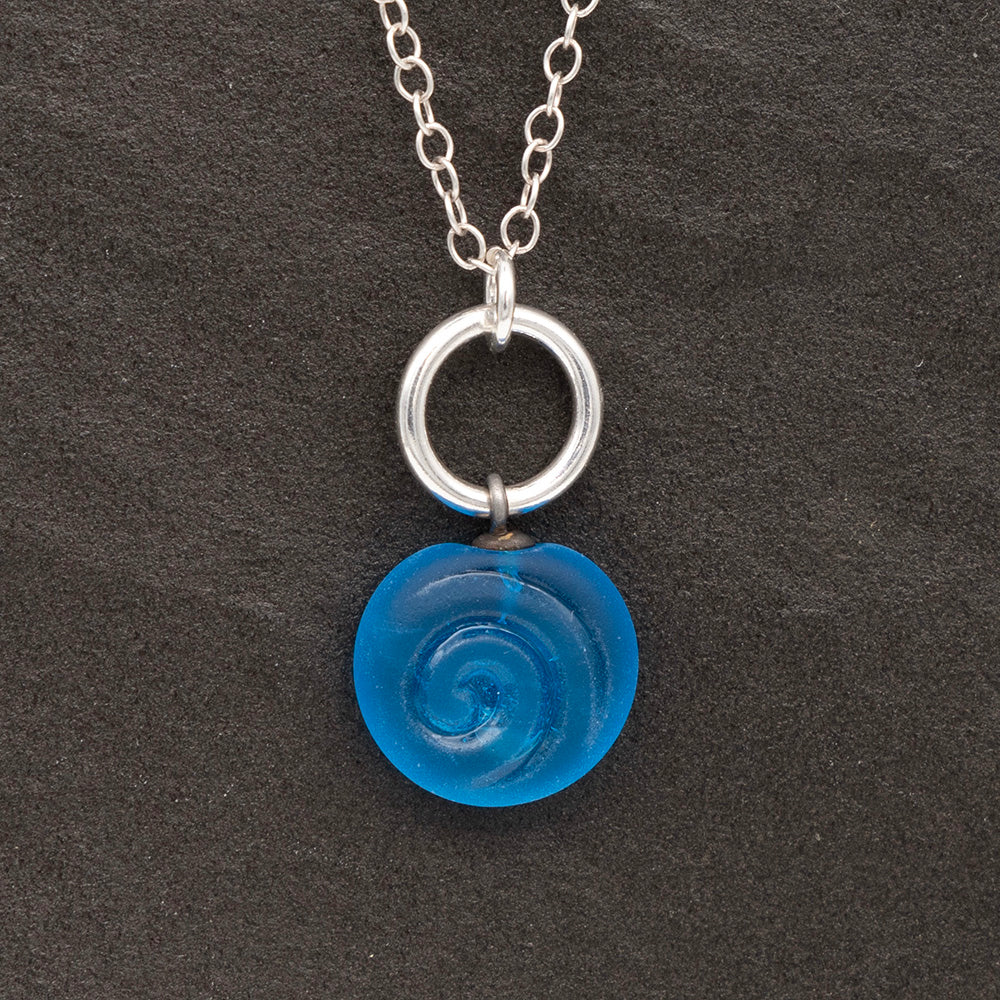 Close up of turquoise frosted glass charm with swirl imprint on a dark slate background. The charm is frosted and spiral is shiny. The charm hangs on a large silver jump ring. The necklace is completed with a small silver jump ring and silver chain.