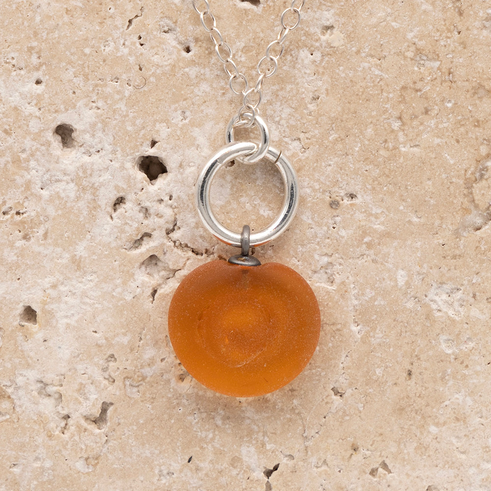 Close up of the back of a yellow frosted glass charm with swirl imprint on a sandstone background. The whole charm is frosted, and spiral is plain frosted glass. The charm hangs on a large silver jump ring. The necklace is completed with a small silver jump ring and silver chain.