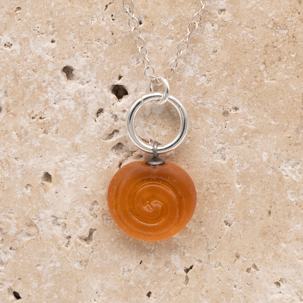 Close up of the front  of a yellow frosted glass charm with swirl imprint on a sandstone background. The charm is frosted and spiral is shiny. The charm hangs on a large silver jump ring. The necklace is completed with a small silver jump ring and silver chain.