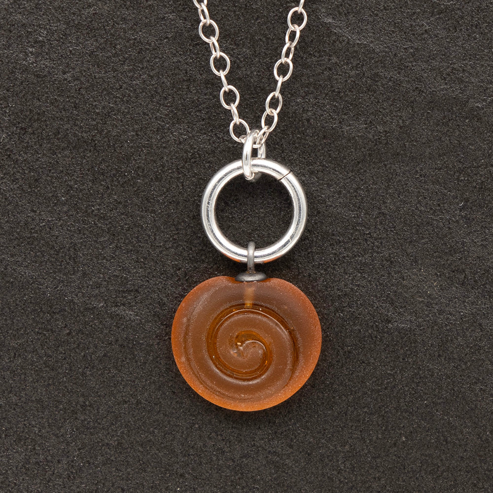 Close up of yellow frosted glass charm with swirl imprint on a dark slate background. The charm is frosted and spiral is shiny. The charm hangs on a large silver jump ring. The necklace is completed with a small silver jump ring and silver chain.