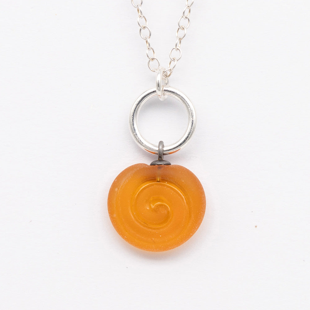 Close up of yellow frosted glass charm with swirl imprint. The charm is frosted and spiral is shiny. The charm hangs on a large silver jump ring. The necklace is completed with a small silver jump ring and silver chain.