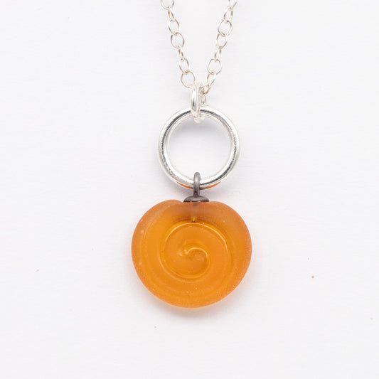 Close up of yellow frosted glass charm with swirl imprint. The charm is frosted and spiral is shiny. The charm hangs on a large silver jump ring. The necklace is completed with a small silver jump ring and silver chain.