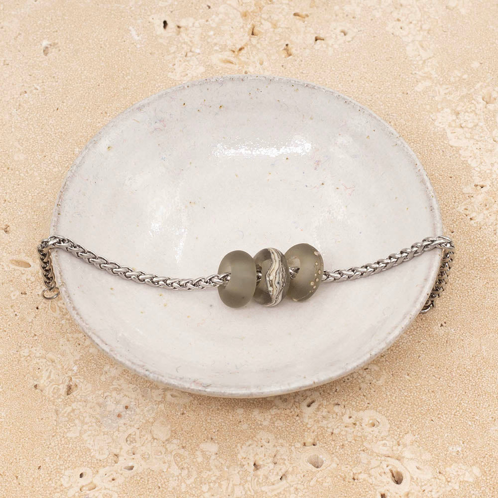 Small white ceramic dish holding three frosted grey glass beads on a stainless steel chain