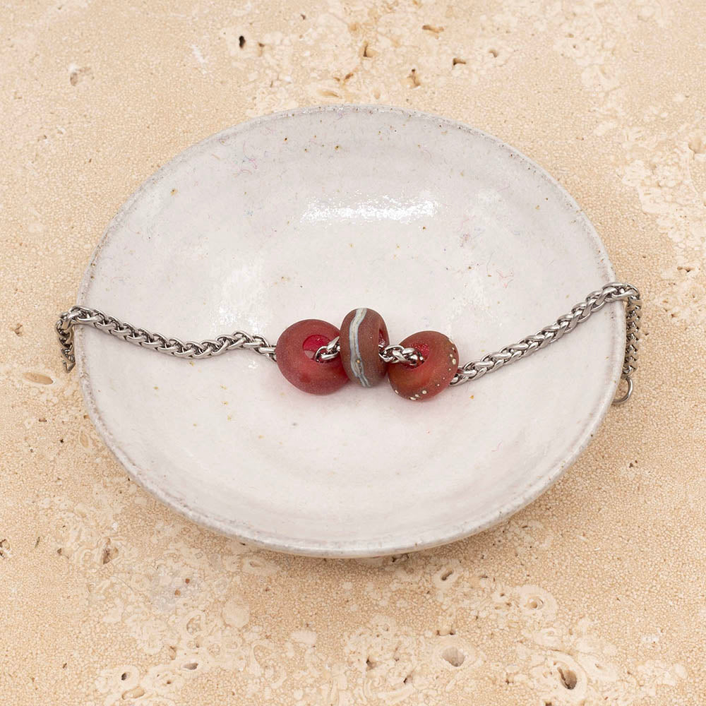 Small white ceramic dish holding three frosted pink glass beads on a stainless steel chain