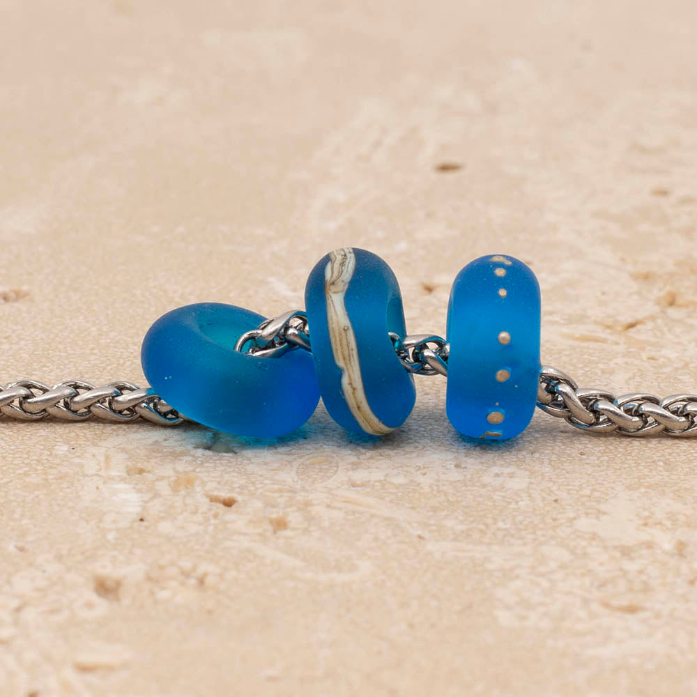 Close up of three frosted turquoise glass beads. One bead is plain, one has a band of ivory and one has tiny silver dots. The beads are on a stainless steel chain.