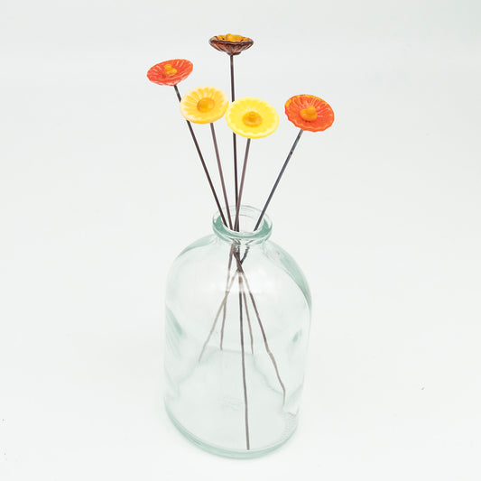 Five yellow and orange glass flowers with metal plant stakes. Flowers are different shades of yellow and orange and have yellow centres.  The plant stakes sit in a clear milk bottle vase.